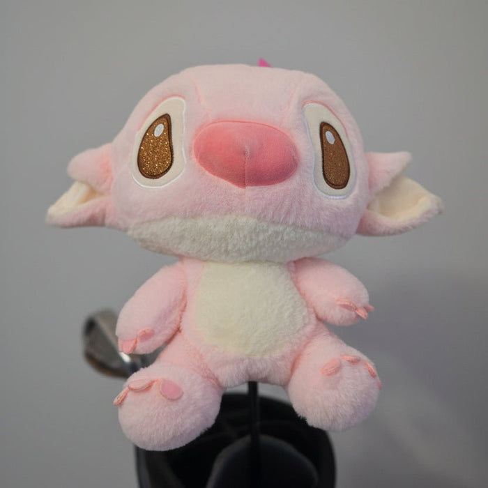 A pink plush toy with large brown eyes and a pink nose, resembling Stitch, sitting on a black stand against a textured beige wall.