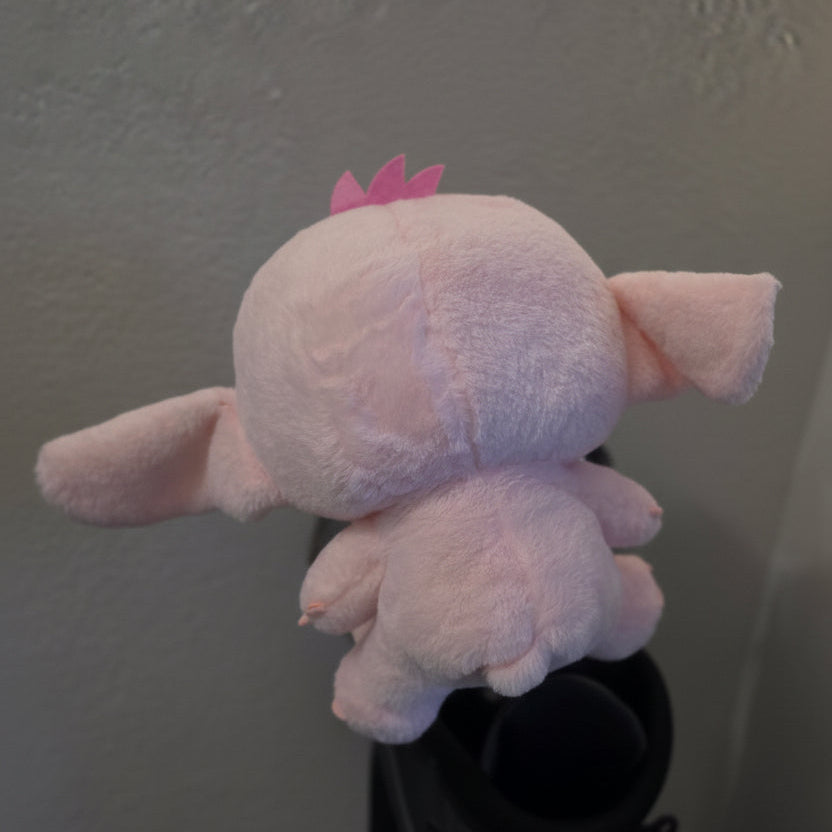 A light pink plush toy with floppy ears and a small pink crown, photographed from the back against a textured off-white wall.