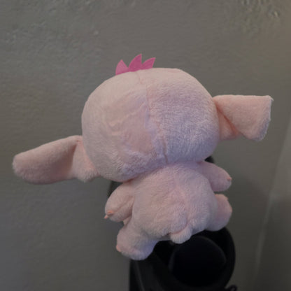 A light pink plush toy with floppy ears and a small pink crown, photographed from the back against a textured off-white wall.