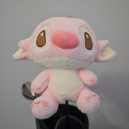 A pink plush toy with large brown eyes and a pink nose, resembling Stitch, sitting on a black stand against a textured beige wall.