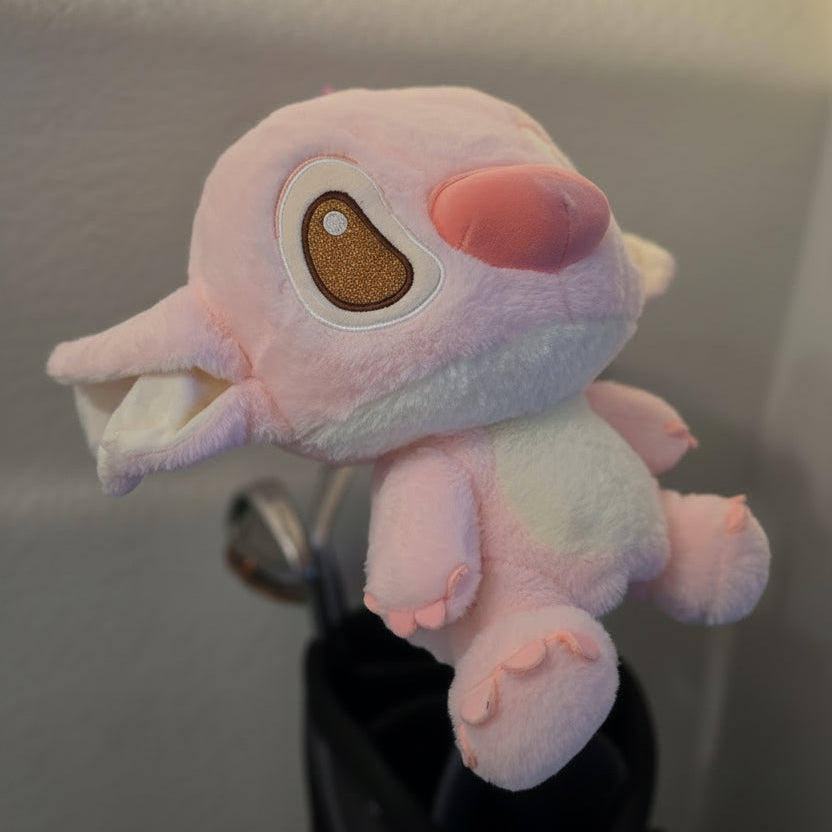 A pink plush toy character with a large embroidered eye and open mouth, resting on top of a black golf bag with clubs visible, against a textured wall.