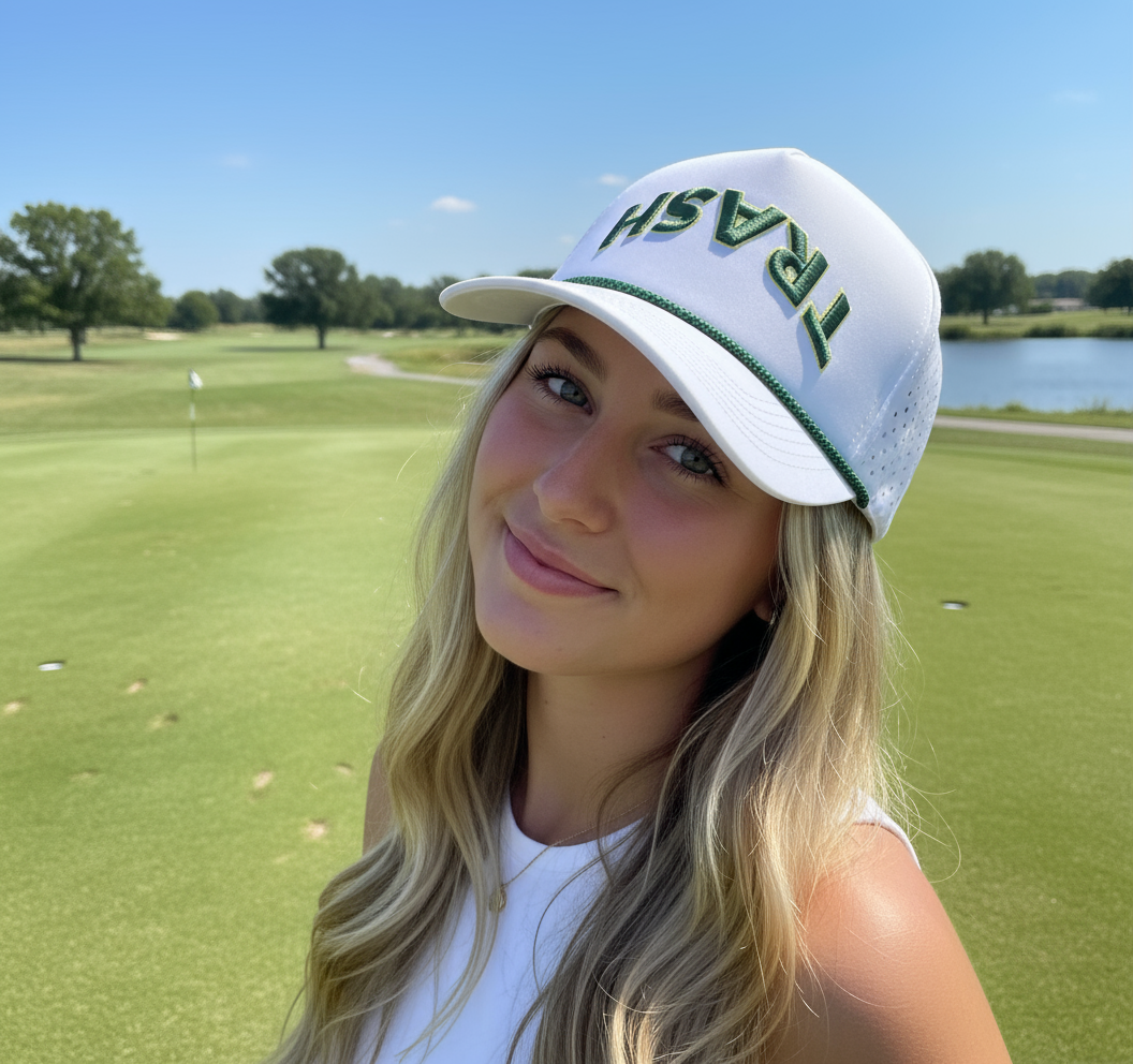 Woman wearing a white cap with 'Trash' printed on it, standing on a golf course.