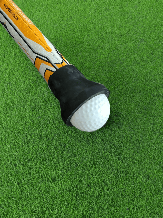 A black rubber golf ball pickup retriever attached to the end of an orange and white putter grip on a green surface, demonstrating the function by lifting a white golf ball.