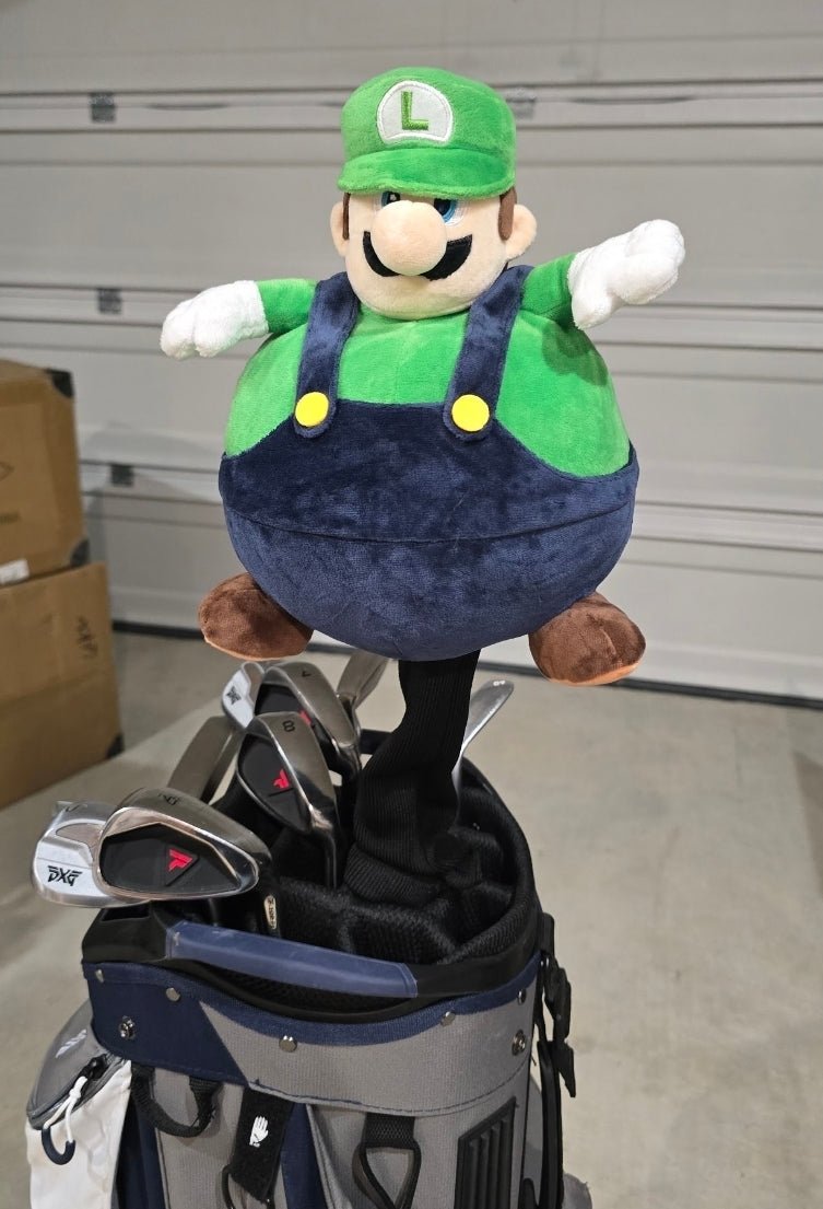 A plush Luigi golf bag topper sitting on top of a navy and gray golf bag with golf clubs inside, photographed in a garage setting.