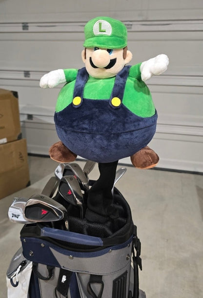 A plush Luigi golf bag topper sitting on top of a navy and gray golf bag with golf clubs inside, photographed in a garage setting.