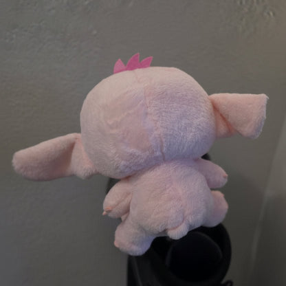 A light pink plush toy with floppy ears and a small pink crown, photographed from the back against a textured off-white wall.
