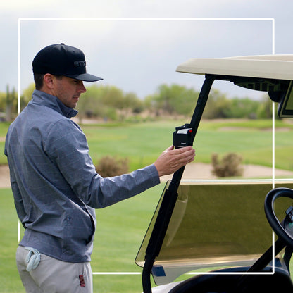 Golfer retrieving a rangefinder with a black STICKIT Gen3 strap from a metal cart post; rangefinder not included