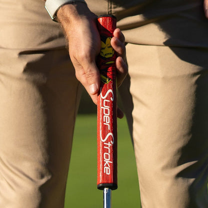 Close-up of a hand holding a red SuperStroke golf putter grip on a blurred golf course background.