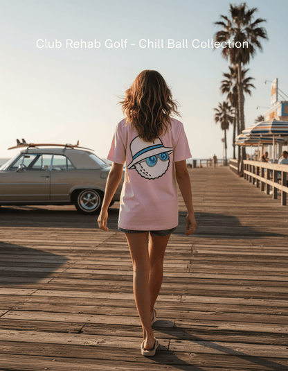 Woman walking on a wooden boardwalk with palm trees and a vintage car in the background, wearing a pink t-shirt with a graphic design.